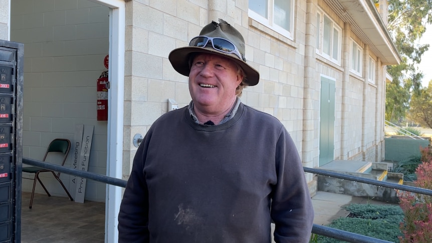 A man smiles, he wears a khaki hat and dark jumper. Behind him is a town hall