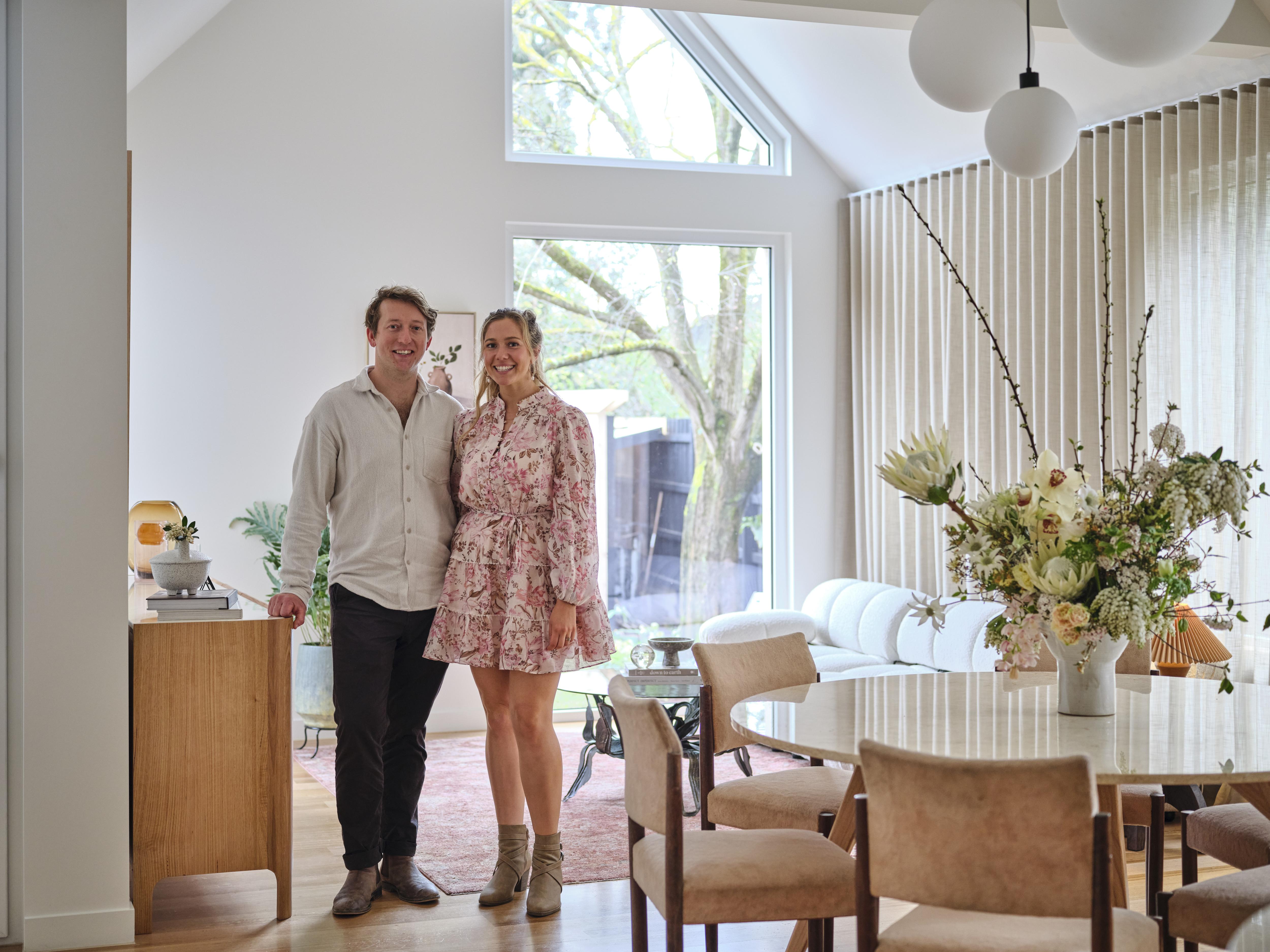 A couple stand in a light filled lounge area with a dining table in the foreground and couch and window in the back