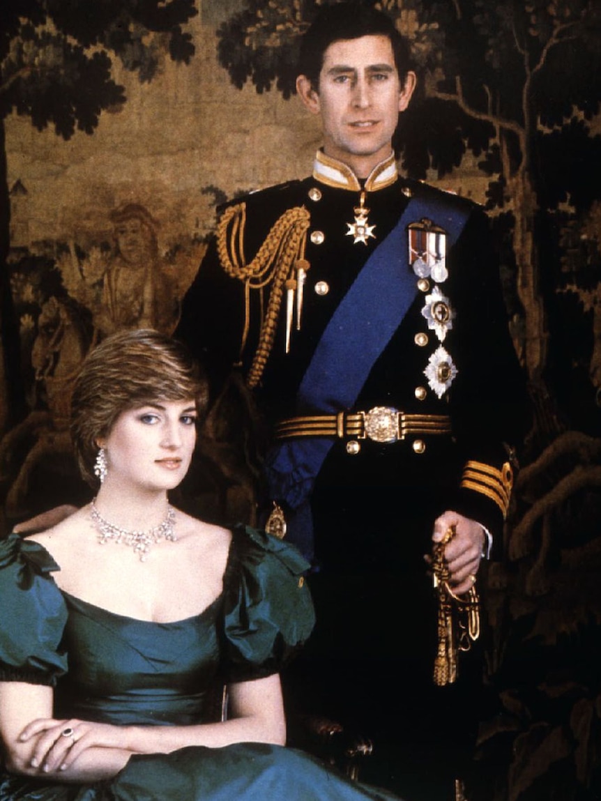 Princess Diana in a deep navy dress sits while Prince Charles stands behind her 