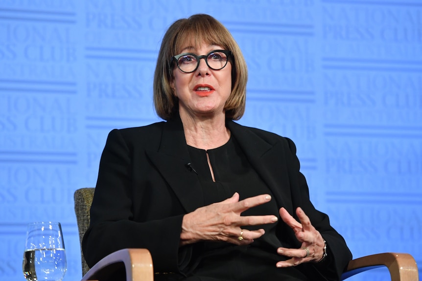 A seated woman wearing glasses and a dark suit talking on stage. She has reddish hair and the background is light blue