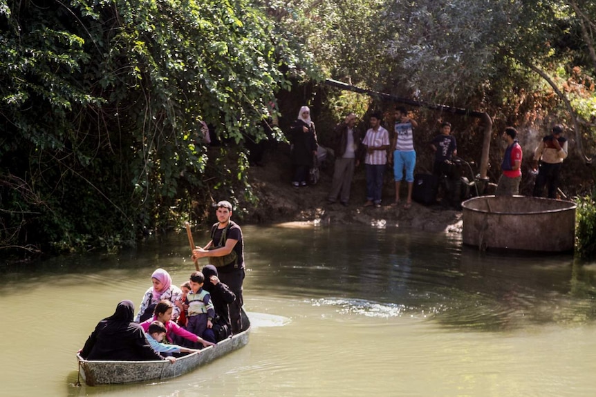 A Syrian family in a tin boat are being paddled across a small, green creek, as people look on from the bank.