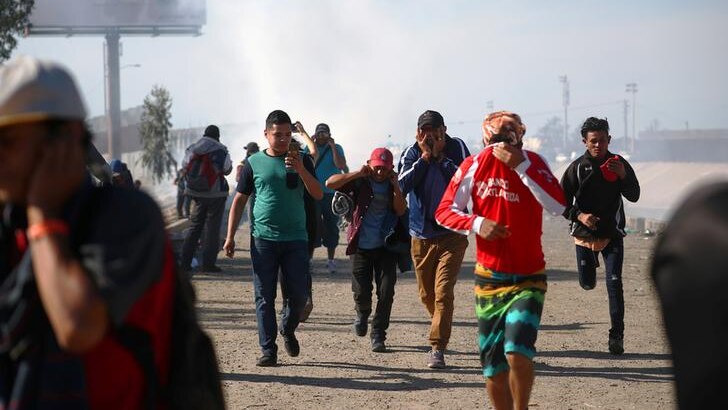 A group of five migrants cover their faces as they flee tear gas in Mexico as gas haze obscures blue skies.