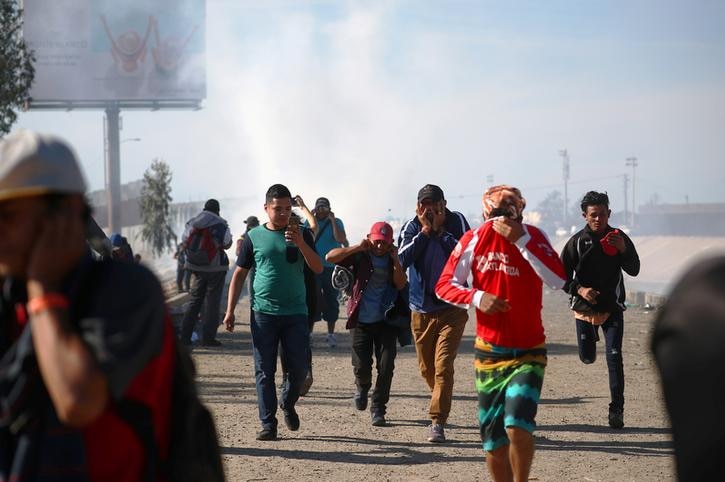 A group of five migrants cover their faces as they flee tear gas in Mexico as gas haze obscures blue skies.