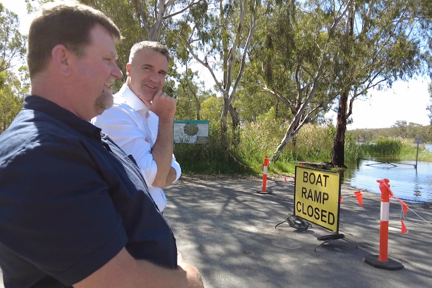 Two men speak next to a river with a sign saying BOAT RAMP CLOSED
