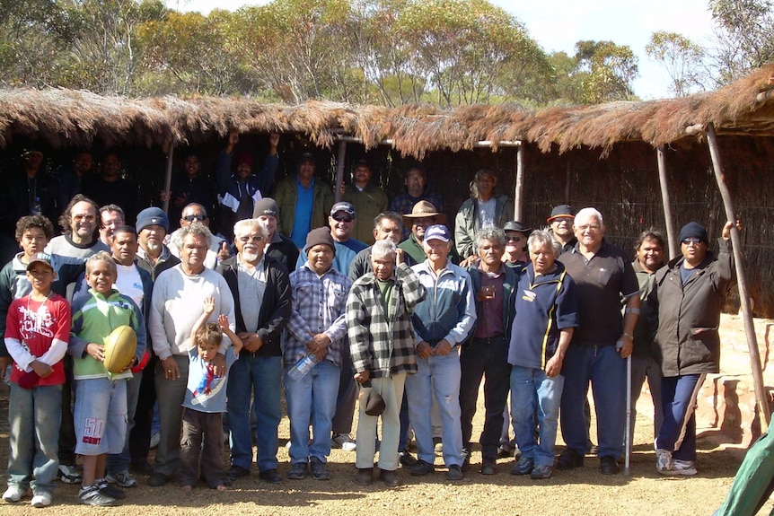 A group of Noongar men outside a pole and thatch shed