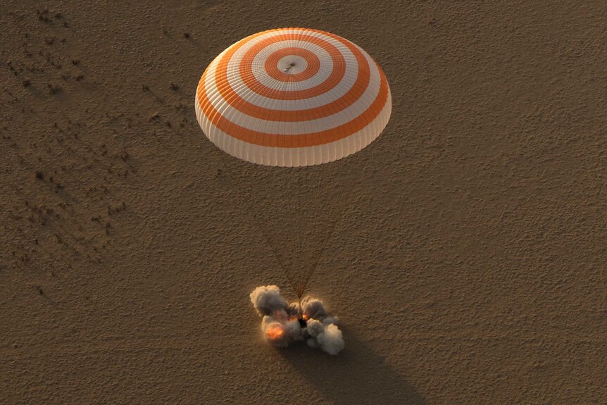 A spacecraft lands in the desert with a puff of sand under a parachute canopy.