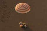 A spacecraft lands in the desert with a puff of sand under a parachute canopy.