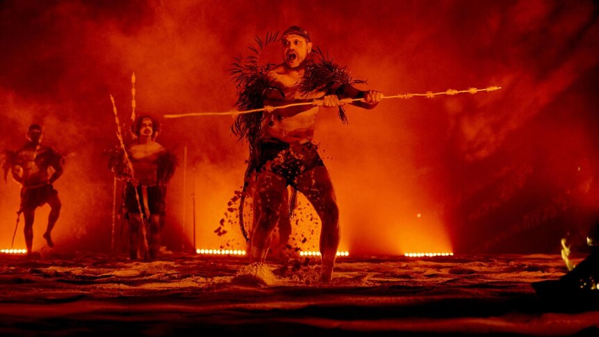 An Indigenous dancer in white face paint holds a spear against a fiery red backdrop/