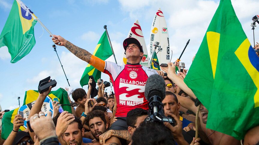 Gabriel Medina, waving a Brazilian flag, sits on the shoulders of many Brazilian fans also holding flags.