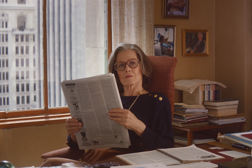 Interior office shot, Sigourney Weave sits at desk dressed stylishly and wearing glasses, while reading paper.