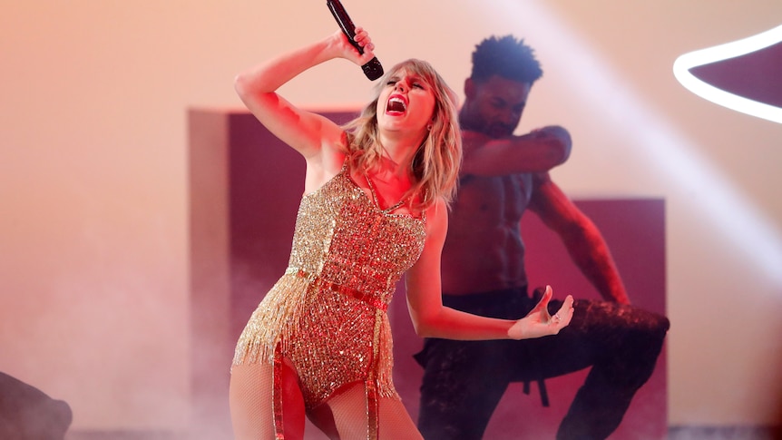 Taylor swift in a gold body suit singing into a microphone with a male dancer behind her
