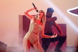 Taylor swift in a gold body suit singing into a microphone with a male dancer behind her