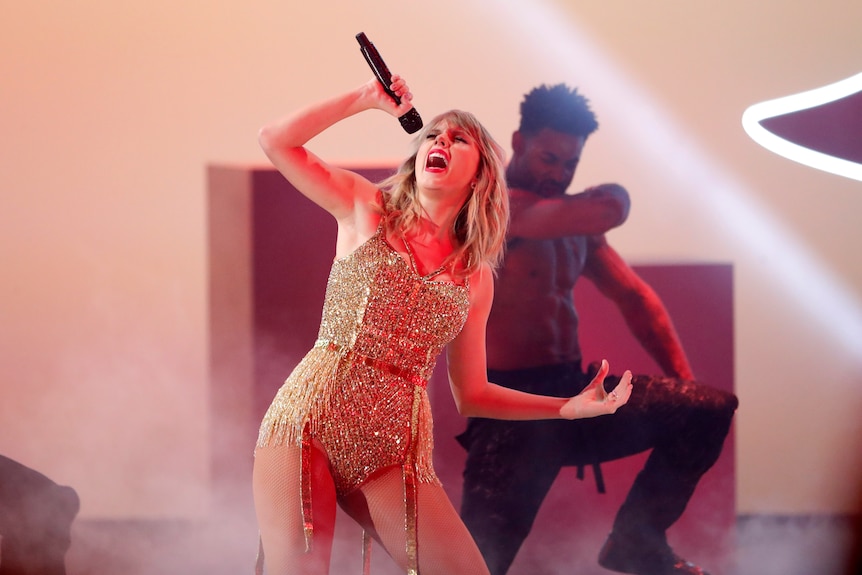 Taylor swift in a gold body suit singing into a microphone with a male dancer behind her