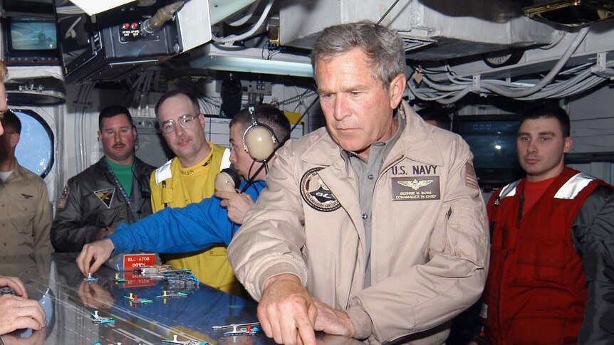 george w bush pointing down on table layout in deck control room wearing us navy coat surrounded by staff and officials