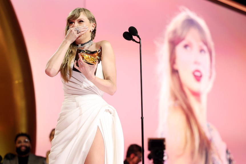 Taylor clasps her hand over her mouth in shock on stage as she holds her award and her face is projected behind her.