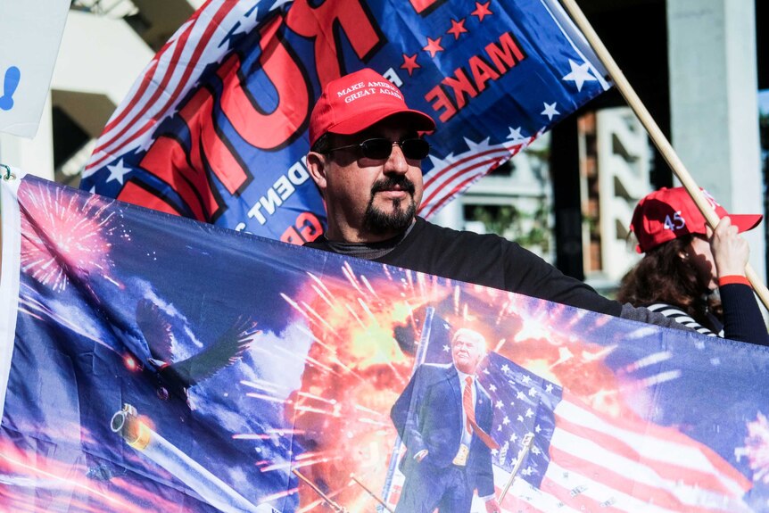 A man holds a flag showing an image of Donald Trump in front of the American flag with an explosion in the background.