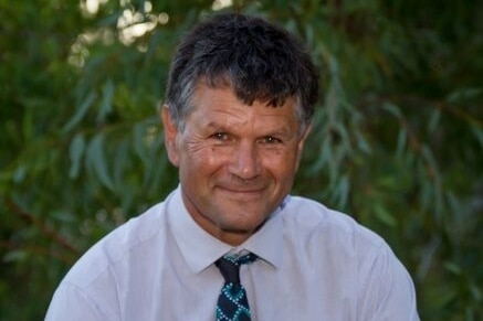 Mid-shot portrait of man in white shirt and tie sitting relaxed smiling at camera with tree behind.