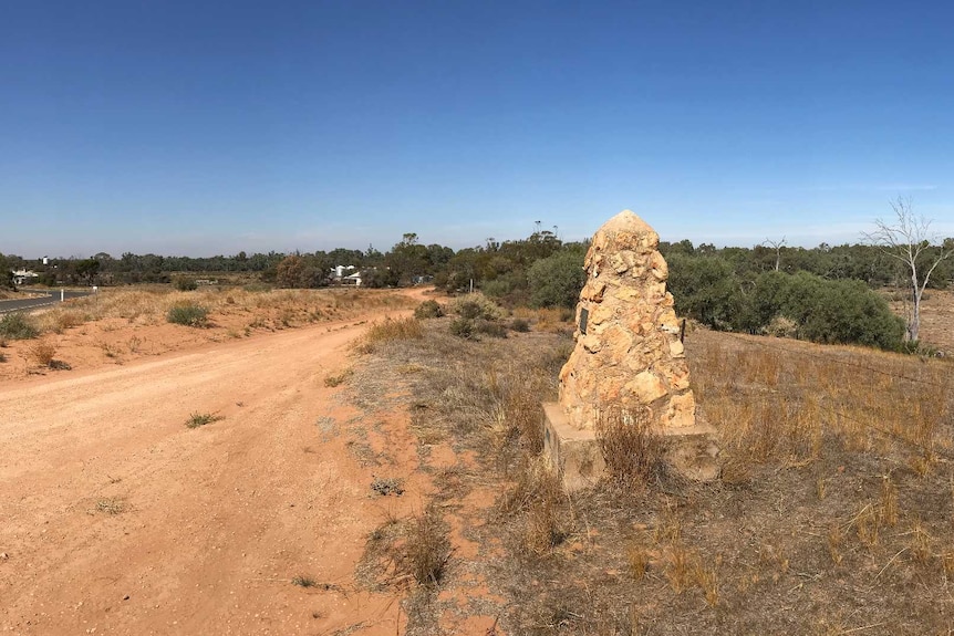A panoramic image of a stone cairn by a roadside with scrub and trees in the background
