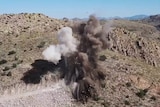 On a clear day, you view an aerial photo of an explosion on a mountainside with dirt and rocks flying into the air.