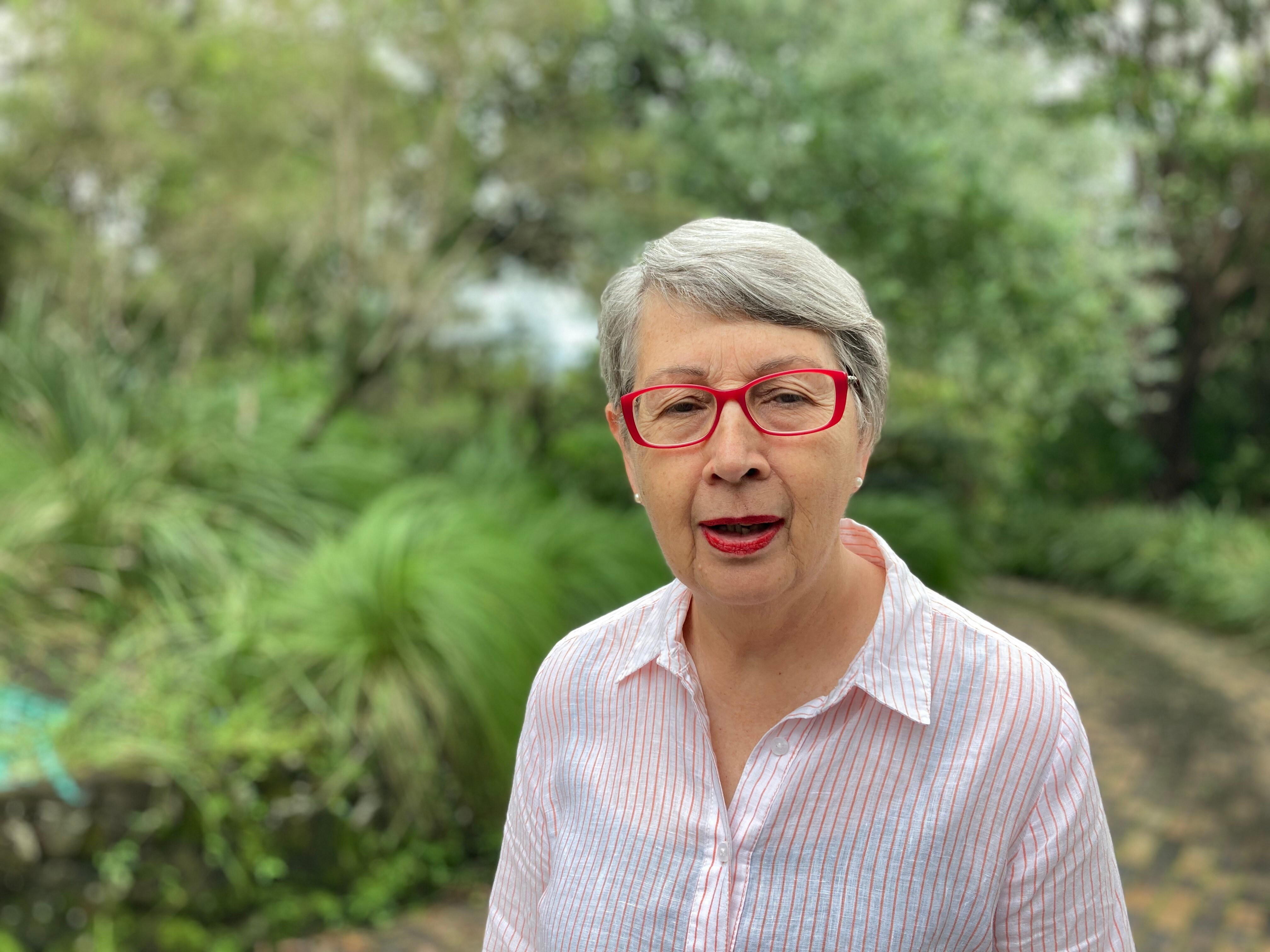 A woman with short grey hair, red glasses and matching red lipstick, wearing a collared button up shirt.