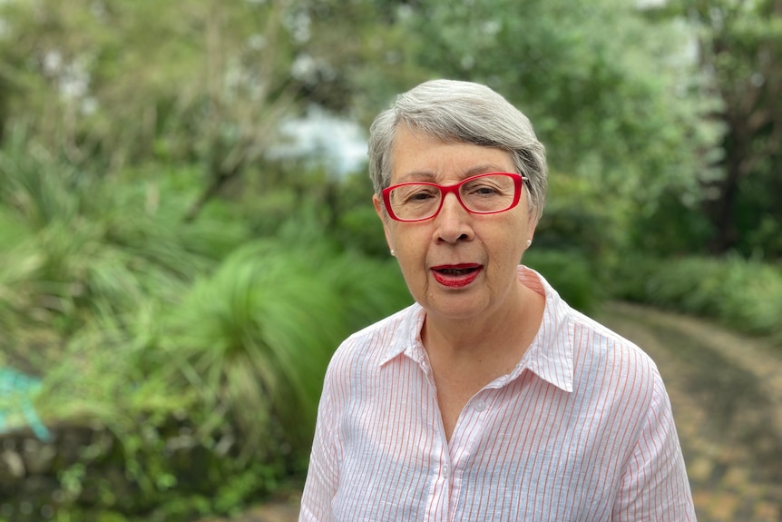 A woman with short gray hair, red glasses and matching red lipstick, wearing a collared button up shirt.