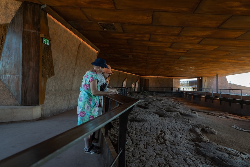Two older women lean on a railing surrounding a length of brown mud with dinosaur footprints, undercover.