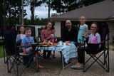 A family, with children, sit on their driveway on chairs, with food set up on a table.