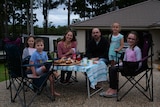 A family, with children, sit on their driveway on chairs, with food set up on a table.