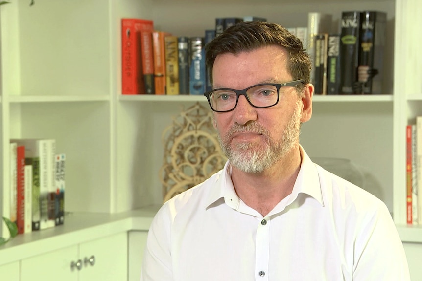 Photo of man with greying beard, wearing glasses and a white shirt in front of a bookcase