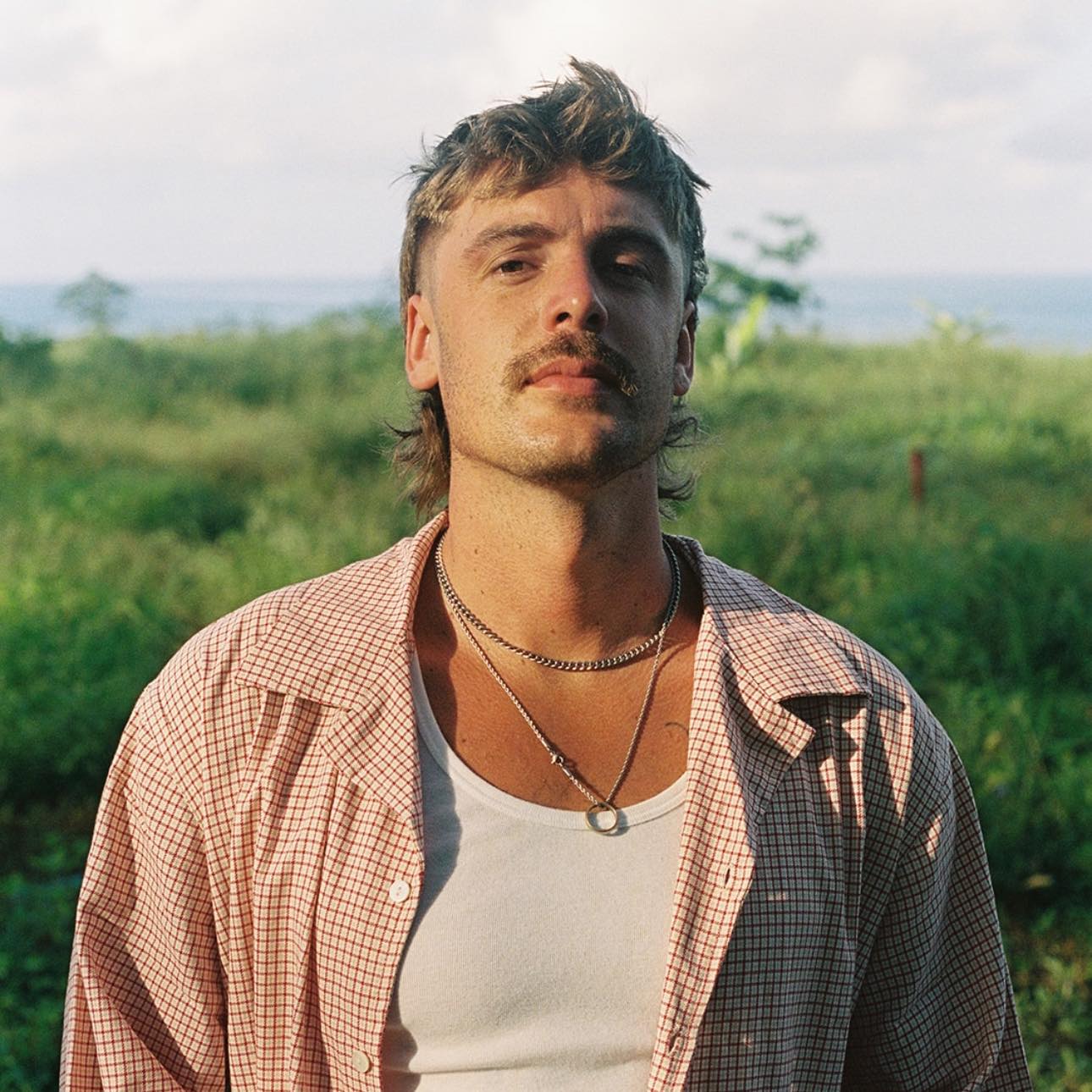 A man in a singlet and open gingham shirt stands against a verdant green field with water on the horizon.