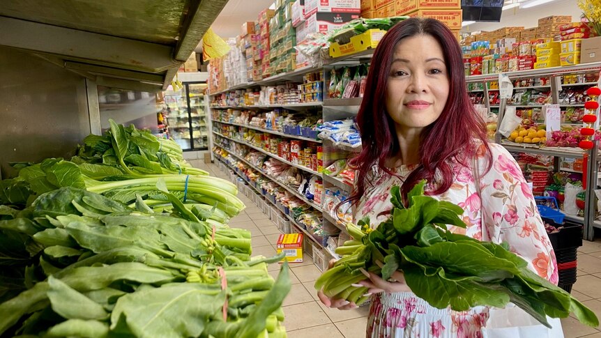 A Vietnamese woman with long red hair holds a leafy green vegetable in the aisle of a grocery store.