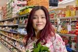 A Vietnamese woman with long red hair holds a leafy green vegetable in the aisle of a grocery store.