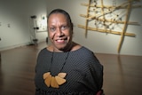 An Aboriginal woman with her hair pulled back smiles at the camera in a low-light art gallery