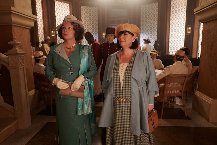 Two prim and proper looking middle-aged women, holding their purses, stand in a crowded sitting room