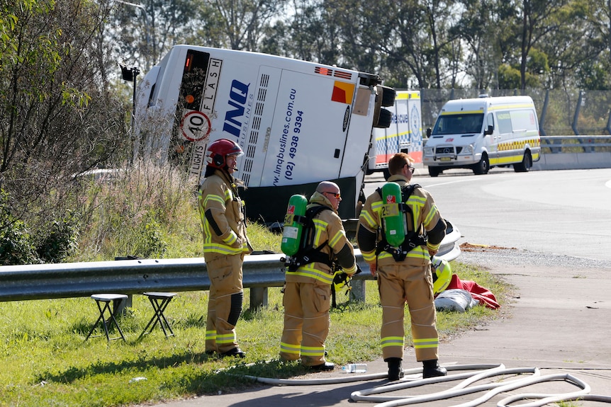 Fire crews stand in front of an overturned bus beside a road.