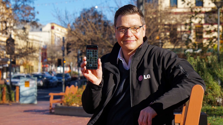 A man in a black jacket holding up a mobile phone while sitting on a park bench.