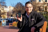 A man in a black jacket holding up a mobile phone while sitting on a park bench.
