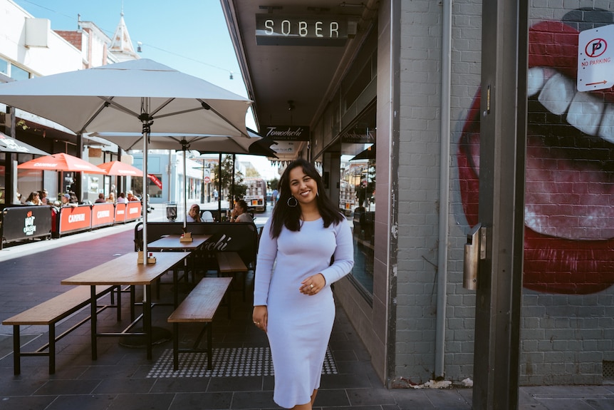 A woman stands in front of a series of bars and restaurants wearing a grey dress. She's under a sober sign, and smiles.