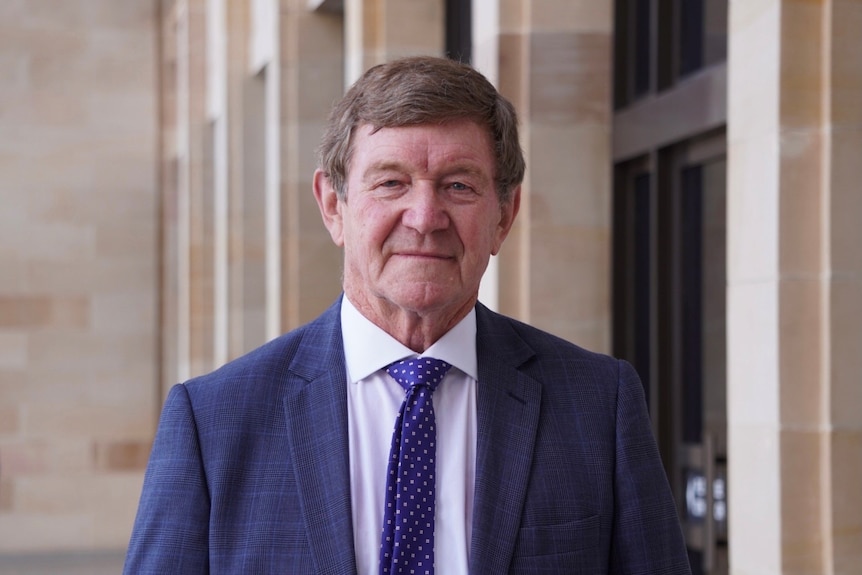 Dr Ron Edwards wears a white shirt and blue suit and tie, standing on the front steps of Parliament House. 