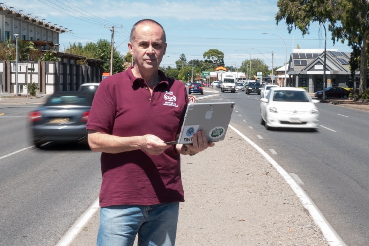 Richard Pascoe holds a laptop, standing on a busy street.