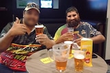 Two men drinking beers, one's face is blurred, he does a thumbs up, the other has a big black beard, smiles.
