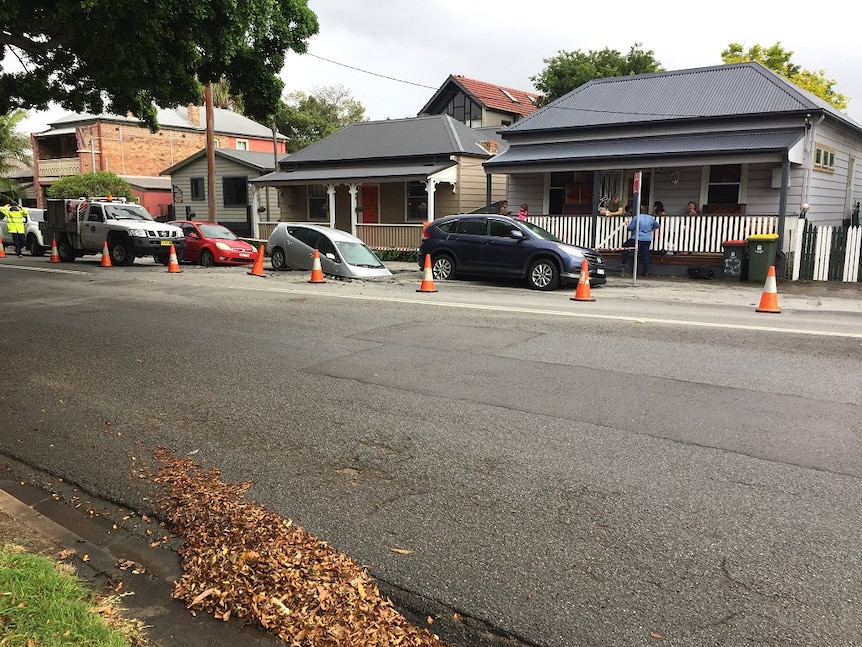 A car rests in a large hole in a roadway, as local residents watch on from nearby homes.