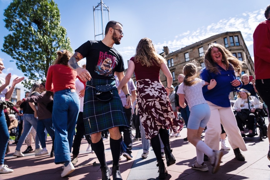 A large number of people, including one wearing a kilt, dancing outdoors.