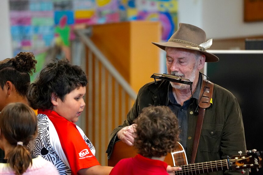 A white man with a brown hat, jacket, guitar and harmonica talking to two young school kids 