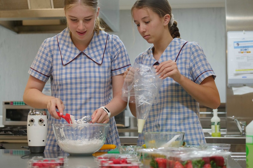 Two teenage girls in school uniform stir food in a bowl to prepare a dessert.
