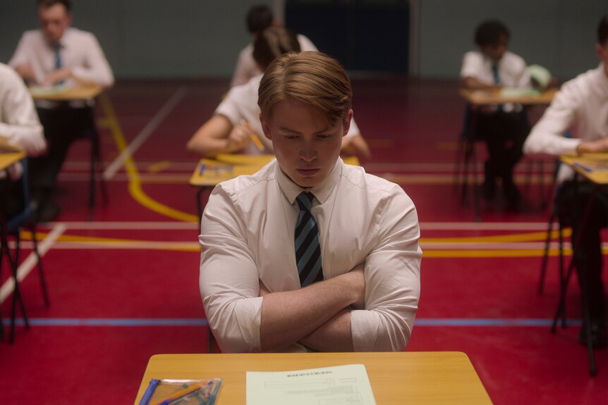 Nick sits at a single school table in school uniform, arms crossed, staring down, looking sad