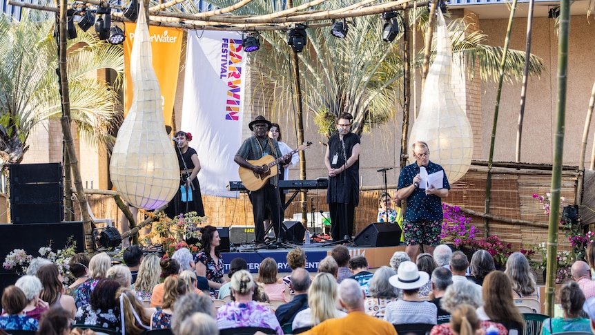 A Mudburra man plays guitar on a bamboo bandstand in the sunshine with backing vocalists and a seated crowd watches