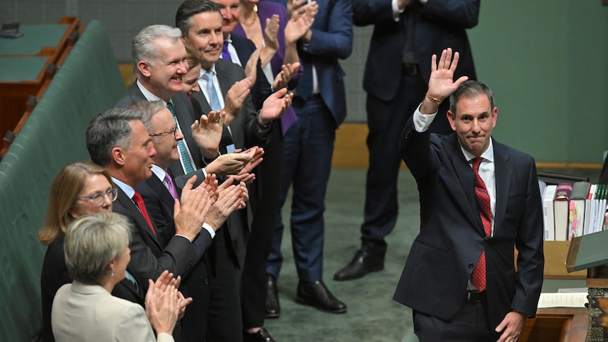 A middle-aged man in a suit waves as he is applauded by a line of people in suits behind him.