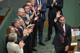 A middle-aged man in a suit waves as he is applauded by a line of people in suits behind him.