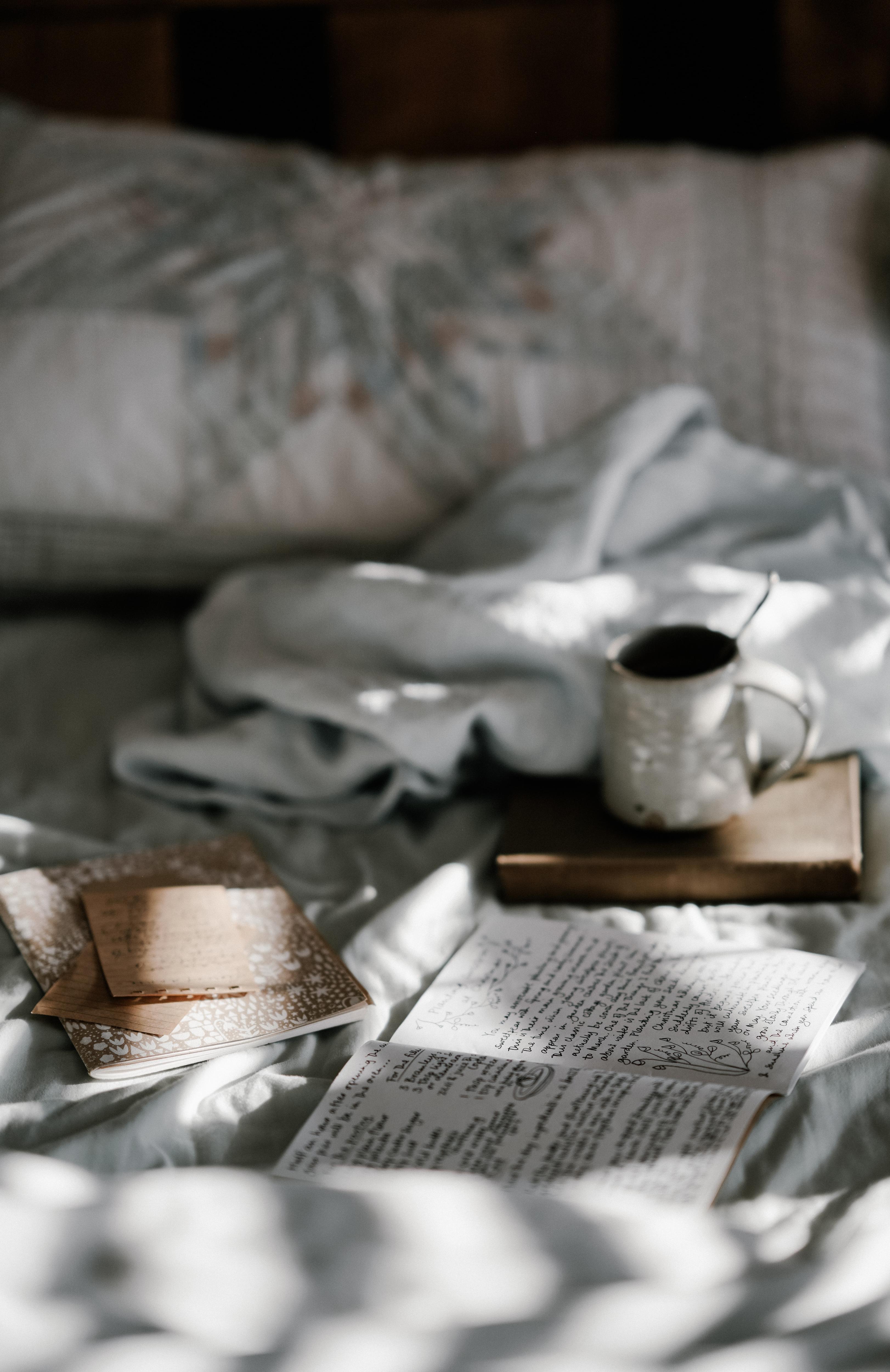 Coffee and notebooks and open journal on bed with white bed linen and dappled morning light.